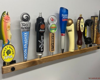 May include: A wooden wall-mounted beer tap handle rack with nine different tap handles. The tap handles feature various designs, including a fish, a pumpkin, a beer can, and a phone. The rack is made of dark wood and has a natural finish. The text "TAP HANDLES NOT INCLUDED" is printed at the bottom of the image.