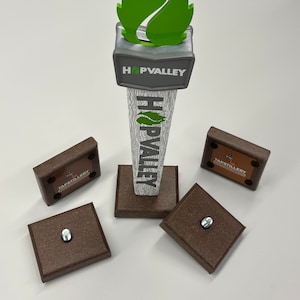 May include: A brown tap handle with a green leaf design and the text "HOPVALLEY" on the front. The tap handle is on a brown base with four smaller brown bases with the text "TAPSTILLERY" on them.