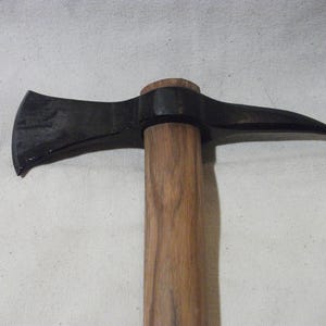 Pode incluir: Um tomahawk com uma cabeça de metal forjado escuro, apresentando uma lâmina afiada e curva de um lado e uma ponta afiada no lado oposto. A cabeça está montada num cabo reto de madeira castanho-clara com textura de grão natural visível.