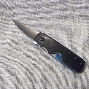 May include: A small, folding pocket knife with a silver blade and a black handle. The knife is open, displaying the sharp blade. The handle has a curved design and is secured with small rivets. The knife is on a textured, light-colored surface.