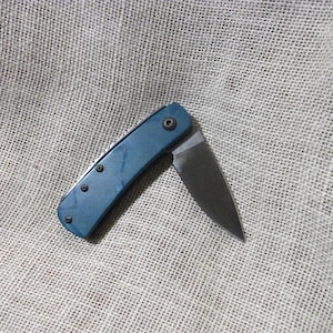 May include: A small, open pocket knife with a teal handle and a silver blade. The handle has three small, dark rivets and a single rivet near the blade. The blade is partially open, revealing its sharp edge. The knife rests on a textured, neutral-colored surface.