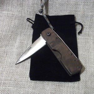 May include: A folding pocket knife with a wooden handle and a silver blade is displayed open, resting on a black velvet pouch. The knife is partially open, revealing the sharp edge. A lanyard is attached to the knife. The background is a textured, off-white fabric.