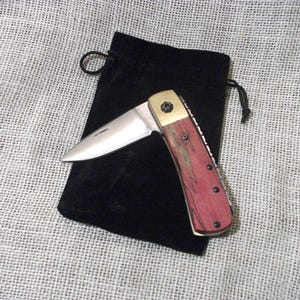May include: A folding pocket knife with a stainless steel blade and a red and brown wooden handle. The knife is open, revealing the blade, and rests on a black velvet pouch. The handle has brass accents.