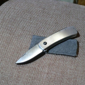 May include: A silver pocket knife with a smooth, curved handle and a sharp blade. The knife is resting on a gray cloth.
