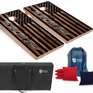 May include: Two wooden cornhole boards with an American flag design and the words "In God We Trust" printed on them. The boards are set up for a game of cornhole, with a black bag and red and blue bean bags in the foreground.