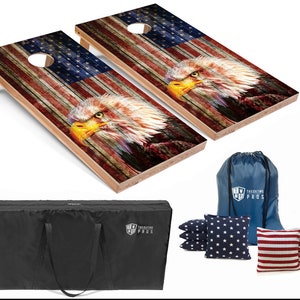 May include: A cornhole game set featuring two wooden boards with an American flag and bald eagle design. Includes bean bags with star and stripe patterns, a carrying bag, and a black storage case. The boards have a hole for tossing bean bags.