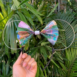 May include: A pair of Minnie Mouse ears with a bow made from a folded map of a theme park. The bow is white with a silver headband.