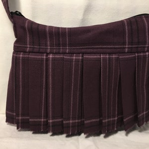 May include: A purple plaid crossbody bag with a pleated bottom and a frayed edge. The bag has a black strap and a gold metal clasp.