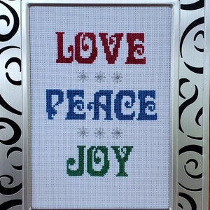 May include: A silver framed cross-stitch with a white background and the words "LOVE PEACE JOY" in red, blue, and green lettering. The frame has a black and silver swirl design.