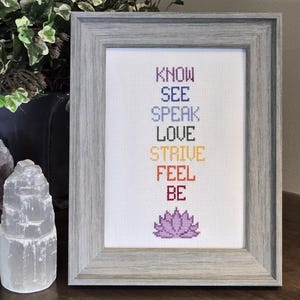 May include: A framed cross-stitch with a gray frame and a white background. The cross-stitch features a rainbow of words: Know, See, Speak, Love, Strive, Feel, Be. A purple lotus flower is at the bottom of the cross-stitch.