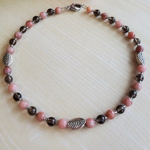May include: A pink and brown beaded necklace with silver leaf charms. The necklace is made with natural stones and is approximately 18 inches long.
