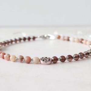 May include: A beaded bracelet featuring a mix of pink, brown, and clear beads. The bracelet has a silver clasp and a decorative metal bead with a floral design. The beads are strung on a clear cord.