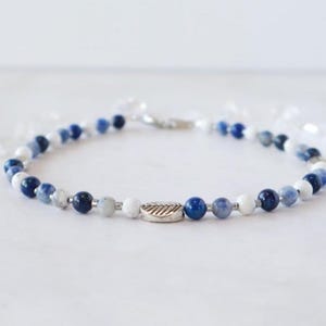 May include: A beaded bracelet featuring a mix of blue, white, and gray beads. A silver-colored leaf charm is centered on the bracelet. The bracelet is strung on a silver-colored chain.