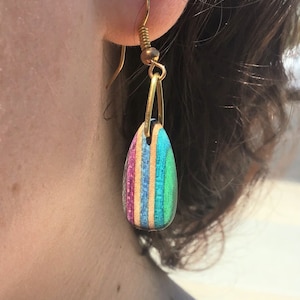 May include: A pair of gold dangle earrings with a teardrop-shaped wooden pendant. The pendant is made of layered wood with a rainbow of colors, including pink, blue, green, and brown.