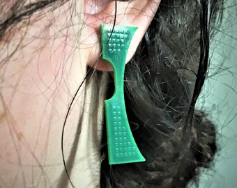 May include: A green plastic earring with a geometric design. The earring is worn in the left ear of a person with dark hair.