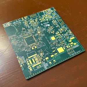 Pode incluir: Uma placa de circuito verde com componentes amarelos e brancos. A placa tem um design complexo com vários componentes eletrônicos e o texto "TALIS". A placa é retangular e está sobre uma superfície marrom.