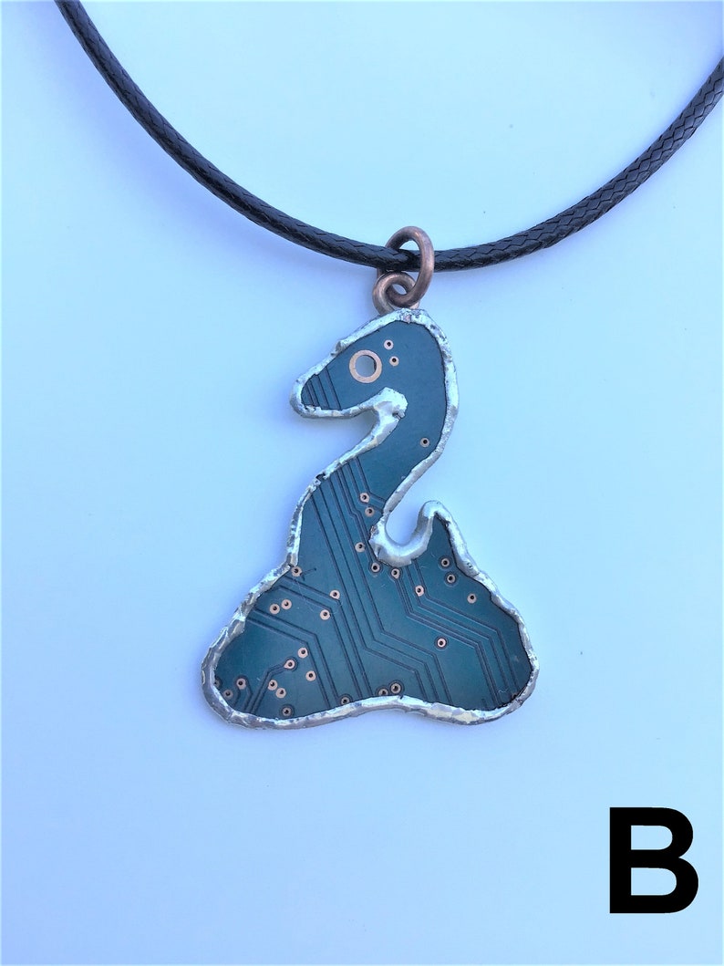 May include: A necklace with a black cord and a pendant shaped like a snake made from a circuit board. The pendant is silver with copper accents and has a green circuit board design.