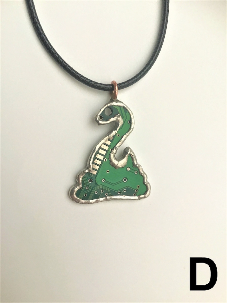 May include: A green circuit board pendant necklace with a snake design. The pendant is attached to a black cord.