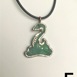 May include: A green circuit board pendant shaped like a snake, with a silver frame and a black cord necklace. The pendant is hanging against a white background.