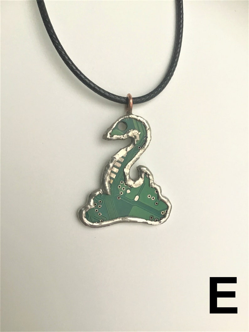May include: A green circuit board pendant shaped like a snake, with a silver frame and a black cord necklace. The pendant is hanging against a white background.