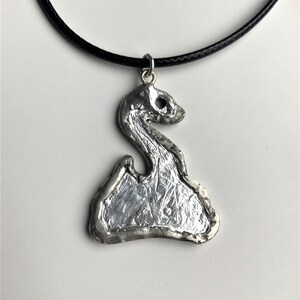 May include: A silver pendant necklace with a textured, abstract design. The pendant is shaped like a stylized swan with a long neck and a rounded body. The necklace is on a black cord.