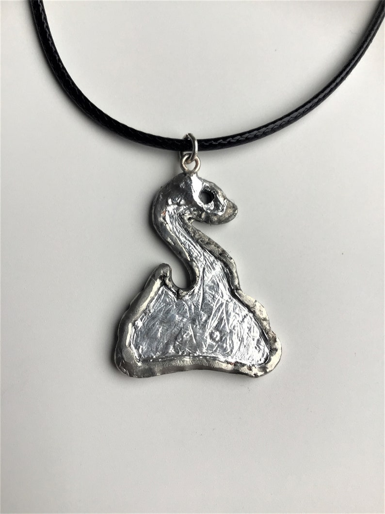 May include: A silver pendant necklace with a textured, abstract design. The pendant is shaped like a stylized swan with a long neck and a rounded body. The necklace is on a black cord.