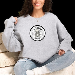 Op de afbeelding: Een lichtgrijs sweatshirt met ronde hals en een cirkelvormige afbeelding op de borst. Het ontwerp bevat een zwart-witte illustratie van een skelet omringd door bloemmotieven, met de tekst LITERALLY DEAD in zwarte hoofdletters gedrukt.