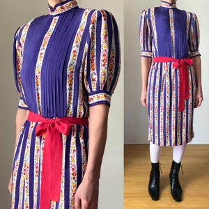 May include: Vintage midi dress with vertical stripes in purple, white, and floral patterns. The dress features a high collar, pleated bodice, short sleeves, and a red ribbon belt. A classic, elegant style.