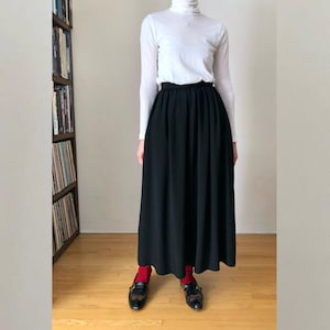 May include: A person wearing a white turtleneck long-sleeve shirt and a long black skirt. The person is wearing red socks and black shoes. A bookshelf filled with books is visible in the background.