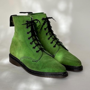 May include: A pair of bright green suede boots with black laces and soles. The boots have a classic lace-up design and a textured suede finish. The boots are a vibrant shade of green, offering a bold and stylish look.