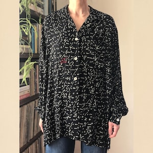 May include: Black button-down shirt with a white mathematical formula print. The long-sleeved shirt has a collared neckline and white buttons. The shirt is worn over blue jeans. A red emblem is visible on the chest.