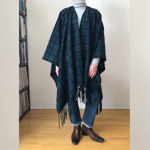 May include: A dark blue and green plaid poncho with fringe. The poncho is worn over a gray turtleneck and blue jeans. Brown leather boots complete the outfit. The background includes a bookshelf and a wooden floor.