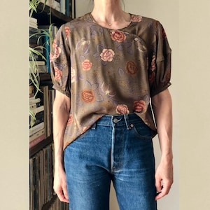 May include: A brown floral print blouse with short sleeves and a round neckline. The blouse features a wrap-style closure with a single button. The model is wearing blue denim jeans. The background includes a bookshelf and a neutral wall.