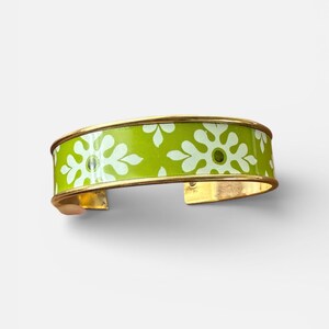 Può includere: Un bracciale a polsino verde lime con un motivo floreale bianco e bordi color oro. Il bracciale aperto è largo circa 5 cm e presenta un motivo floreale ripetuto.