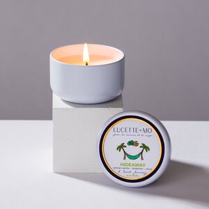 May include: A white candle in a tin container with a label that reads "LUCETTE+MO pour la maison et le corps" and "HIDEAWAY tahitian vanilla cardamon citrus A Scent Journey 6 oz Candle".