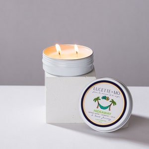 May include: A white tin candle with a blue and green label that reads "LUCETTE+MO pour la maison et le corps HIDEAWAY Tahitian vanilla, cardamom, citrus A Scent Journey Love Travel Tin 2 oz". The candle is lit and has two wicks.