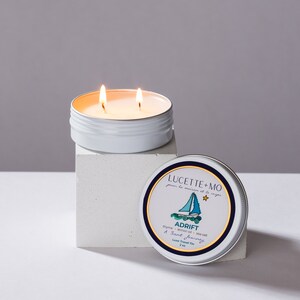 May include: A white tin candle with two wicks burning. The candle is labeled "Lucette + Mo" and "Adrift" with the text "A Scent Journey" and "Luxe Travel Tin 2 oz".