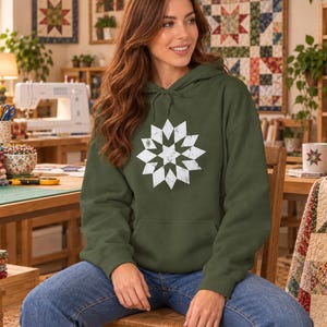 May include: An olive green hooded sweatshirt featuring a white, distressed-style geometric star quilt block graphic on the chest. The hoodie includes a front kangaroo pocket, long sleeves, and matching olive green drawstrings at the neckline.