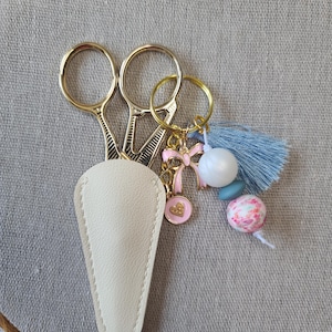 May include: Gold-toned embroidery scissors with a cream-colored leather sheath. A decorative keychain with a pink bow, heart charm, blue tassel, and floral bead is attached. The scissors are on a neutral linen background.