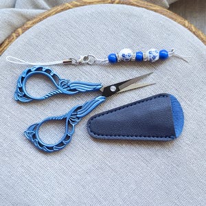 May include: Blue embroidery scissors with a matching leather sheath and a beaded charm. The scissors have a decorative handle and sharp blades. The charm features blue and white beads. The items are displayed on a light gray fabric.