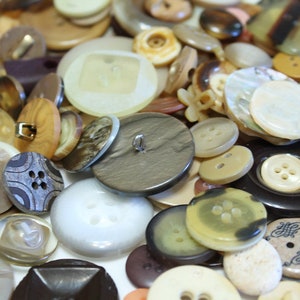 May include: A collection of assorted buttons in various colors, shapes, and sizes. The buttons are made of different materials, including plastic, metal, and wood. Some buttons have a shiny finish, while others have a matte finish.