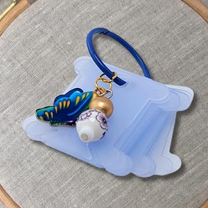 May include: A blue plastic thread bobbin holder with a gold and blue butterfly charm and a white and gold bead. The bobbin holder is on a light brown fabric surface.