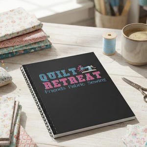 Puede incluir: Un cuaderno negro con espiral con el texto "QUILT RETREAT Friends Fabric Sewing" en verde azulado y rosa. El cuaderno está sobre una superficie de madera con tela, una taza de café, tijeras y una bobina de hilo.