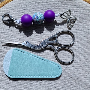 May include: A collection of craft supplies including a pair of silver embroidery scissors with a bird design, a light blue leatherette scissor sheath, and a beaded charm with purple and blue beads and a butterfly charm.
