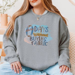May include: A gray sweatshirt with the text "zero days without buying fabric" in colorful, stylized lettering. The text is set against a watercolor-style background of blue, pink, and yellow hues. The sweatshirt is a casual garment.