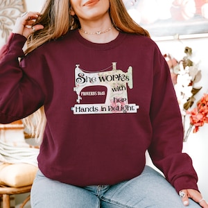 Peut inclure: Sweat-shirt bordeaux avec un motif de machine à coudre et le texte "She works with her Hands in Delight" et "Proverbes 31:13". Le haut à manches longues est fait d'une matière douce.