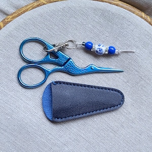 May include: Metallic blue stork-shaped embroidery scissors with a decorative charm featuring blue and white beads attached to the handle, paired with a dark blue leather sheath. The scissors have a detailed textured finish on the metal surface.