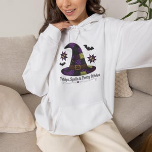 May include: A white hooded sweatshirt featuring a graphic of a purple, green, and black patchwork witch hat with a brown belt buckle. The design includes two star shapes, two bats, and the text Patches, Spells & Pretty Stitches in black font.