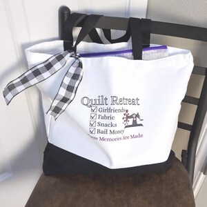 May include: White tote bag with black accents and handles, featuring the text "Quilt Retreat" and a list of essentials. A black and white checkered ribbon is tied to the handle. The bag is sitting on a brown chair.