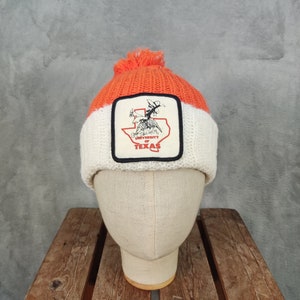 May include: Orange, white, and black knitted beanie hat with a pom-pom. The hat has a patch with the text "UNIVERSITY OF TEXAS" and a graphic of a person riding a horse. The beanie is displayed on a mannequin head.